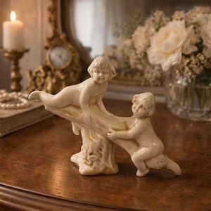 Könnte beinhalten: Eine cremefarbene Figur, die zwei cherubische Figuren zeigt, die auf einem Ast spielen. Die Skulptur steht auf einer dunklen Holzoberfläche, mit einer brennenden Kerze und einer Vase mit Blumen im Hintergrund.