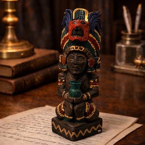 May include: A carved wooden statue of a figure with a headdress and a mask. The statue is brown with red, blue, green, and gold accents. The figure is holding a green vase. The statue is sitting on a piece of paper.