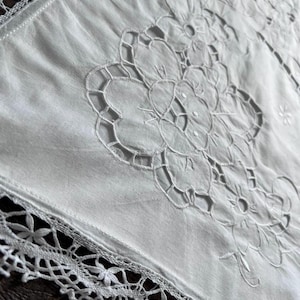 May include: A white rectangular textile featuring intricate cutwork embroidery in a floral pattern. The edges are finished with a delicate, wide lace border that displays a repeating star-like motif, creating a decorative and detailed fabric piece.
