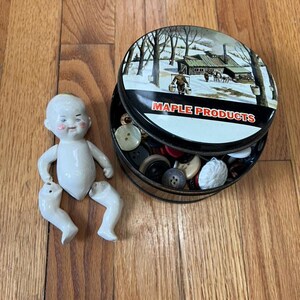 May include: A white porcelain doll with a detached torso and legs, positioned next to a round black metal tin labeled MAPLE PRODUCTS. The tin lid features a winter scene with a cabin and trees, and the container is filled with assorted buttons.