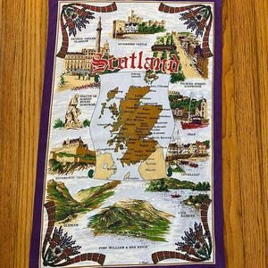 May include: A rectangular tea towel with a purple border featuring a map of Scotland in the center. The design includes illustrations of Inverness Castle, Edinburgh Castle, Glencoe, and Loch Katrine, with the word Scotland printed in red.