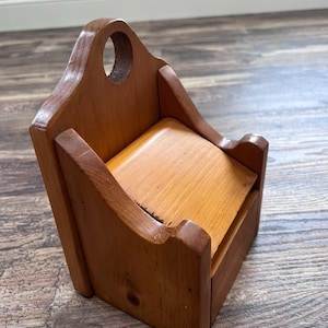 May include: A wall-mounted wooden salt box with a hinged lid and a circular cutout at the top for hanging. The container features a medium-brown wood grain finish, a slanted front panel, and high side walls that taper toward the front edge.