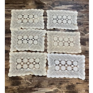May include: A set of six rectangular cream-colored lace doilies featuring a central pattern of circular motifs surrounded by a diagonal grid design and finished with a scalloped edge. Each piece displays intricate needlework and openwork details.
