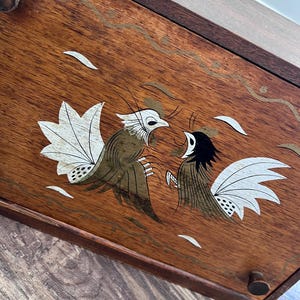 May include: A rectangular wooden bread box featuring a dark brown finish and a hinged front door. The front panel is decorated with a painted illustration of two stylized birds facing each other, rendered in shades of white, black, and gold.