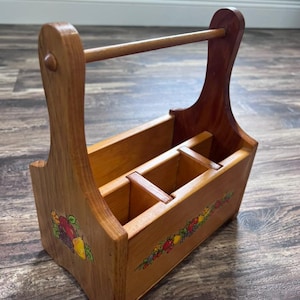 May include: A wooden caddy with a central handle and three interior compartments. The exterior features painted fruit illustrations, including apples, grapes, and pears, on the side and front panels, showcasing a natural wood grain finish throughout.