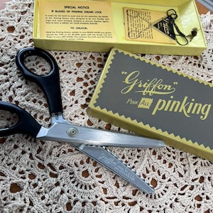 May include: A pair of silver-toned metal pinking shears with black plastic handles, accompanied by a yellow box labeled Griffon Pink All Pinking, an instruction card, and a small triangular tag with black string, all featuring printed text.