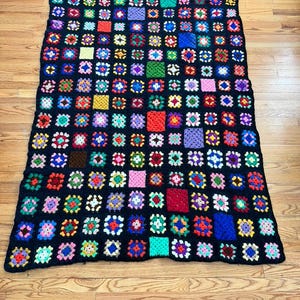 May include: A handmade crochet blanket featuring a patchwork design of colorful squares on a black background. The squares are in various colors, including red, blue, green, and purple. The blanket is rectangular and appears to be made of yarn.