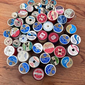 May include: A large collection of assorted sewing thread spools in various colors, including red, blue, white, and green. Each spool features a circular label with brand names like Coats & Clark, Talon, and Singer, along with text indicating thread type, yardage, and price.