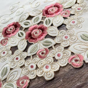 May include: A cream-colored fabric runner featuring intricate cutwork embroidery. The design showcases deep pink roses, light pink rosebuds, and sage green leaves outlined in gold thread, accented by small embroidered floral motifs.