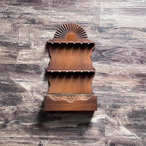 May include: A brown wooden wall-mounted display rack featuring a semi-circular sunburst carved top, two horizontal shelves with six small notches each for holding items, and a solid rectangular base compartment with a textured metal inlay.