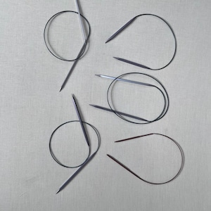 May include: A collection of four circular knitting needles with metallic silver-toned tips and flexible black cords. The needles are arranged in a scattered pattern, showcasing the pointed ends and the circular structure of the connecting cables.