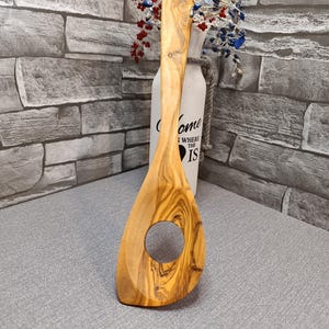 Könnte beinhalten: Ein Kochlöffel aus Olivenholz mit einem großen kreisförmigen Loch in der Mitte. Der Löffel hat einen langen Griff und ein natürliches Holzmaserungsmuster. Es ist ein Küchenutensil.