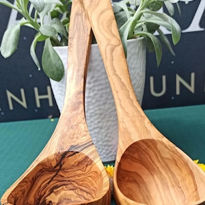 Può includere: Due cucchiai di legno d'ulivo con un motivo a venature naturali. I cucchiai hanno una coppa profonda e un lungo manico con un foro. Sono esposti davanti a un vaso bianco con piante verdi.