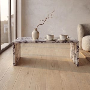 May include: A rectangular coffee table featuring a white and dark grey marble pattern with a waterfall edge design. On the table sits a cream-coloured ceramic vase containing a single thin branch and two white ceramic cups on matching saucers.