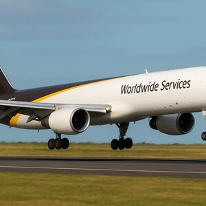 May include: A UPS Boeing 757-200F cargo aircraft in flight, with the UPS logo on the tail and the words "Worldwide Services" on the fuselage. The plane is white with brown and yellow accents, landing on a runway.