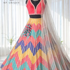Puede incluir: Un lehenga multicolor sobre un maniquí. La falda presenta un estampado de chevrón en amarillo, rosa, azul y turquesa, con detalles dorados. La parte superior es de color coral, con cuello en V, y una dupatta blanca transparente está drapeada sobre el hombro. El logotipo de la marca "NP UNIQUE" es visible.