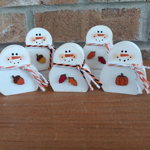 May include: Five white wooden snowman figurines with painted black eyes, orange carrot noses, and orange and black scarves. Each snowman has a different autumn-themed embellishment: an acorn, leaves, or a pumpkin. The snowmen are on a wooden surface.