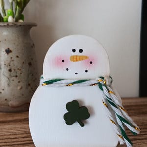 May include: A white wooden snowman figurine with a green shamrock on its front. The snowman has a carrot nose, black dot eyes, and rosy cheeks. A green and white scarf with gold accents is around its neck.