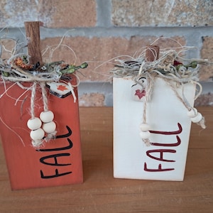 May include: Two rectangular wooden blocks shaped like pumpkins, one orange and one white, each featuring the word FALL in black or dark red lettering. They are topped with cinnamon stick stems, dried raffia, and small wooden beads on twine.