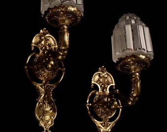 May include: Two antique wall sconces with a gold finish. Each sconce features a clear, rectangular glass shade and ornate, decorative detailing. The sconces are mounted on a black background, highlighting their intricate design and vintage appeal.