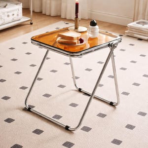 Könnte beinhalten: Kleiner, zusammenklappbarer Tisch im Retro-Stil mit einer transparenten, orangefarbenen Tischplatte und Chrombeinen. Auf dem Tisch stehen Toast, eine Kerze und ein kleiner Behälter. Der Tisch steht auf einem gemusterten Teppich.
