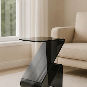 Op de afbeelding: Een moderne zwarte bijzettafel met een uniek Z-vormig ontwerp. De tafel heeft een glanzende afwerking en een rechthoekig blad. Hij staat op een neutraal gekleurd tapijt bij een raam en een beige bank.