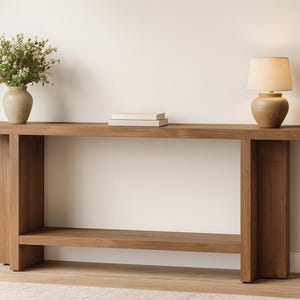 Könnte beinhalten: Eine braune Holzkonsolentisch mit unterem Regal, auf dem eine Vase mit Grün, eine kleine Lampe und zwei Bücher stehen. Der Tisch hat ein minimalistisches Design und eignet sich für Wohnzimmer oder Eingangsbereiche.