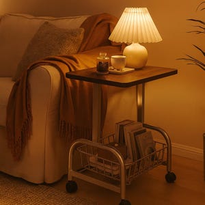 May include: A small, square side table with a dark wood top and a metal frame. A white lamp with a pleated shade sits on the table, along with a cup and a small bottle. A metal basket on wheels holds books.