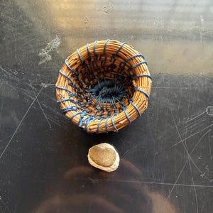 Peut inclure: Un petit panier rond tressé à la main, fabriqué à partir d'aiguilles de pin séchées et lié avec du fil bleu, accompagné d'un galet unique, lisse et brun clair, placé directement sous le panier sur une surface sombre et réfléchissante.