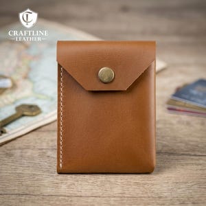 May include: A tan leather card holder featuring a triangular flap closure secured with a single brass-toned metal snap button. The wallet has visible white stitching along the left side and a smooth, matte finish on the front surface.