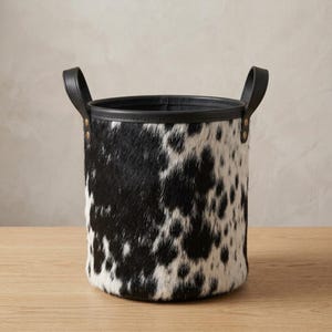 May include: A cylindrical storage basket featuring a black and white cowhide pattern with a textured fur finish. The basket includes a solid black leather-like rim and two matching black handles secured with gold-toned metal rivets on each side.
