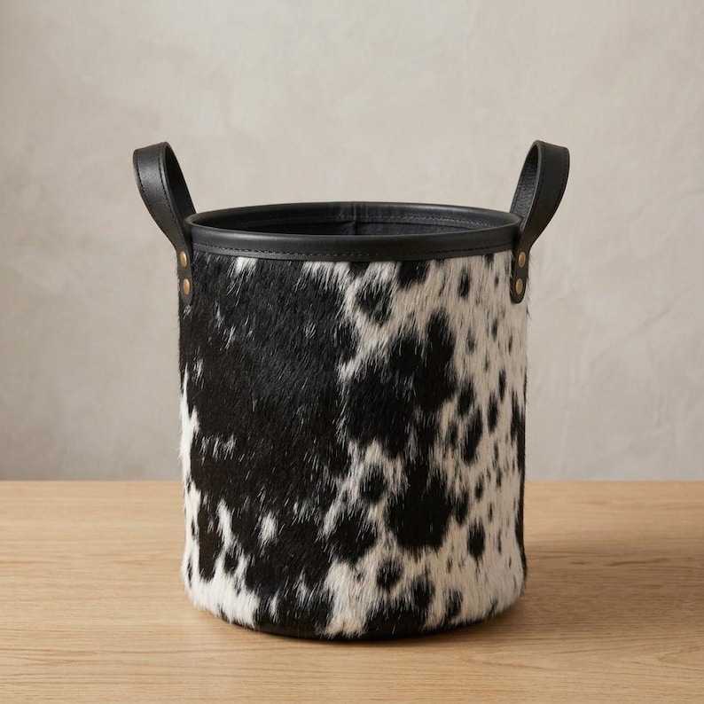 May include: A cylindrical storage basket featuring a black and white cowhide pattern with a textured fur finish. The basket includes a solid black leather-like rim and two matching black handles secured with gold-toned metal rivets on each side.