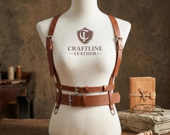 May include: A tan leather harness featuring two vertical shoulder straps with silver-toned buckles, connected to a horizontal waist belt and a lower strap. The chest area displays the text CRAFTLINE LEATHER with a shield logo above it.