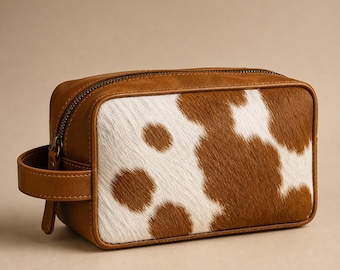 May include: A rectangular toiletry bag featuring a brown and white cow print pattern on the front panel. The sides, top, and handle are made of smooth brown material. A metal zipper runs along the top edge, and a small handle is attached to the side.