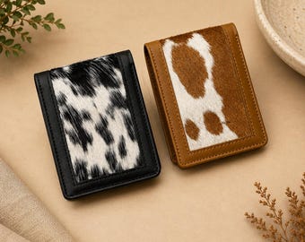 May include: Two rectangular wallets featuring cowhide panels. The left wallet has black and white fur, while the right wallet has brown and white fur. Both wallets are framed with solid leather borders in matching black and tan colors.