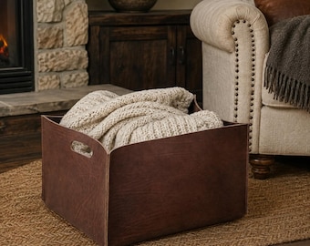 May include: A square, dark brown leather storage bin with integrated side handles. The bin is filled with a thick, cream-colored, chunky knit blanket that spills over the top edges, creating a soft contrast against the smooth leather surface.