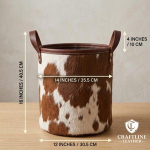 May include: A cylindrical storage basket featuring a brown and white cowhide pattern with a smooth brown leather rim and two side handles. Dimensions are 16 inches high, 14 inches wide, 12 inches at the base, and 4 inches for the handle height.