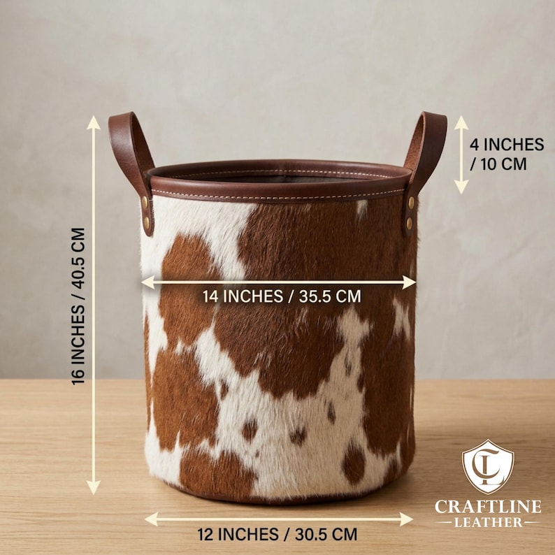 May include: A cylindrical storage basket featuring a brown and white cowhide pattern with a smooth brown leather rim and two side handles. Dimensions are 16 inches high, 14 inches wide, 12 inches at the base, and 4 inches for the handle height.