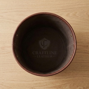 May include: A round, dark brown leather container with visible stitching along the rim and sides. The interior base features a stamped logo with a shield emblem and the text CRAFTLINE LEATHER. The structure is held together by small metal rivets.