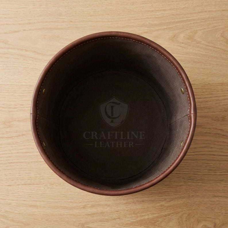 May include: A round, dark brown leather container with visible stitching along the rim and sides. The interior base features a stamped logo with a shield emblem and the text CRAFTLINE LEATHER. The structure is held together by small metal rivets.