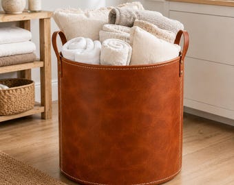 May include: A cylindrical storage basket made of smooth, cognac-colored faux leather with visible white stitching along the seams and two sturdy side handles. The basket is filled with several folded white and beige fabric towels and pillows.