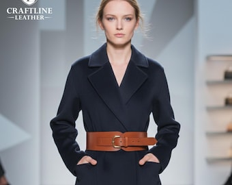 May include: A woman wearing a navy blue coat with a wide, brown leather belt. The belt has a gold-colored buckle. The Craftline Leather logo is in the upper left corner. The coat has a classic design with a notched collar and pockets.
