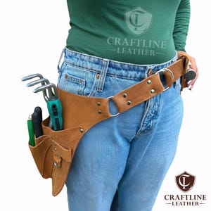 Op de afbeelding: Een bruine leren tuingereedschapsriem met meerdere zakken en een metalen ring. De riem wordt om de taille gedragen en bevat diverse tuingereedschappen. Het Craftline Leather-logo is zichtbaar.
