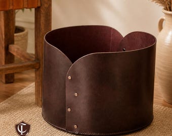 May include: A dark brown leather storage basket with a curved top and brass rivets. The interior is a deep burgundy color. The basket is cylindrical and sits on a woven rug. The Craftline Leather logo is visible.