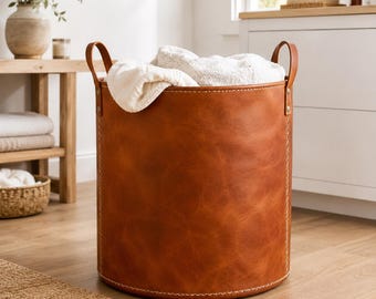 May include: A cylindrical storage basket crafted from smooth, cognac-colored leather featuring visible light-colored stitching along the seams and two sturdy leather handles attached with metal rivets, filled with soft, white folded fabric.