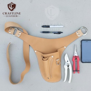 Op de afbeelding: Een bruinleren tuingereedschapsriem met meerdere zakken en een riem. De riem bevat een witte en rode snoeischaar, een smartphone en twee pennen. Het logo "CRAFTLINE - LEATHER" is zichtbaar.
