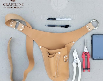 May include: A tan leather gardening tool belt with multiple pockets and a strap. The belt holds a pair of white and red pruning shears, a smartphone, and two pens. The logo "CRAFTLINE - LEATHER" is visible.