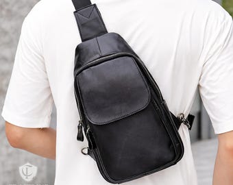 May include: A black leather sling bag with a zippered main compartment and a flap pocket. The bag has a shoulder strap and is worn across the body. The bag is made of leather and has a minimalist design.