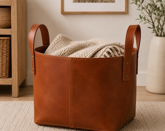 May include: A round, cognac-colored storage basket made of smooth material with two tall, vertical handles attached by metal rivets. The basket features a central vertical seam and contains a folded, textured, cream-colored knit blanket.