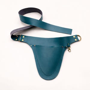 Op de afbeelding: Teal leren schort met een lange riem en een zak. De schort heeft messing klinknagels en een messing ring. De riem is grijs aan de onderkant. De schort is ontworpen om om de taille te worden gedragen.